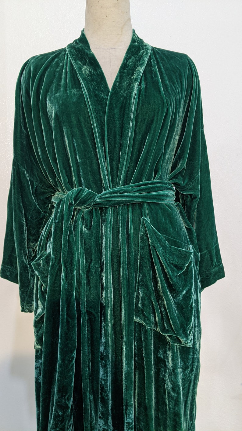 May include: A long, dark green velvet robe with a shawl collar and a tie belt. The robe has two patch pockets on the front.