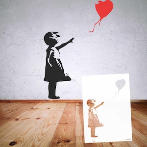 May include: Black silhouette of a girl reaching for a red heart-shaped balloon on a white wall. A stencil of the same image is on a white card. The artwork is on a wooden floor.