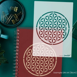 May include: A white stencil with a geometric flower of life design. The stencil is on a green surface with a red notebook and gold paper clips. The text "Example DIN A4 (18cm)" is at the bottom of the image.
