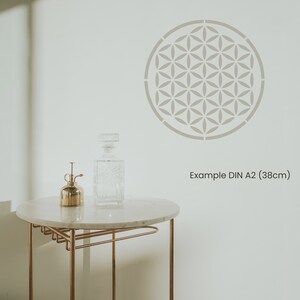 May include: A white wall with a gray geometric stencil design in the shape of a circle. The stencil is 38 cm in diameter and is labeled "Example DIN A2 (38cm)". A small gold metal spray bottle and a clear glass bottle are on a white marble table with a gold metal frame.