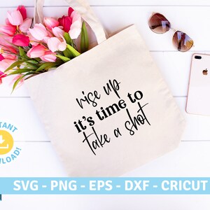 May include: A white tote bag with the text "Rise up it's time to take a shot" in black script font. The bag is lying on a white wooden surface with a bouquet of pink tulips and a pair of sunglasses.