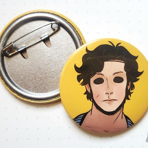 May include: A yellow button with a black and white illustration of a person with black hair and a white mask. The person is wearing a blue and white striped shirt.