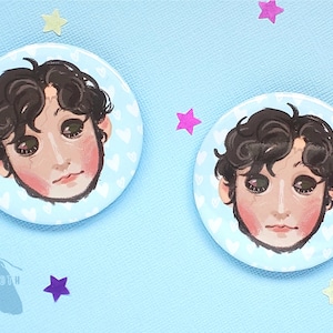 May include: Two round button pins with a blue background and white hearts. Each pin features a cartoon portrait of a person with brown hair and green eyes.