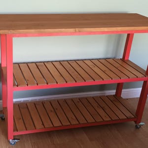 May include: A wooden kitchen island with a natural wood top and slatted shelves. The frame is painted a bright red, and it has wheels for easy movement. The island is approximately 91 cm wide, 61 cm deep, and 91 cm tall.