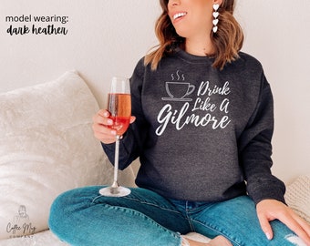 Könnte beinhalten: Dunkelgraues Sweatshirt mit dem weißen Schriftzug "Drink Like A Gilmore". Das Design zeigt eine Kaffeetasse und Untertasse. Das Model trägt das Sweatshirt mit blauen Jeans und hält eine Champagnerflöte.