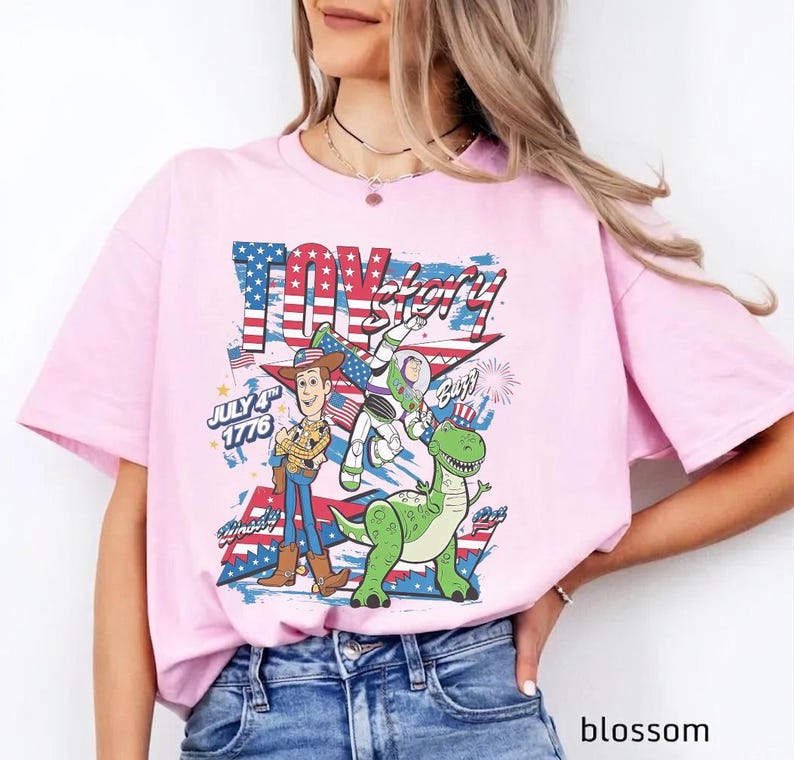 Op de afbeelding: Lichtroze t-shirt met een grafisch ontwerp uit de film "Toy Story". Het ontwerp bevat Woody, Buzz Lightyear en Rex, met de woorden "Toy Story" en "July 4th 1776" in een patriottisch kleurenschema.