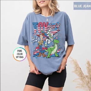 Op de afbeelding: Lichtblauw T-shirt met een Toy Story-afbeelding. Het toont Woody, Buzz Lightyear en een groene dinosaurus, met de tekst "Toy Story" en "July 4th 1776". Het ontwerp bevat elementen van de Amerikaanse vlag. Korte mouwen.