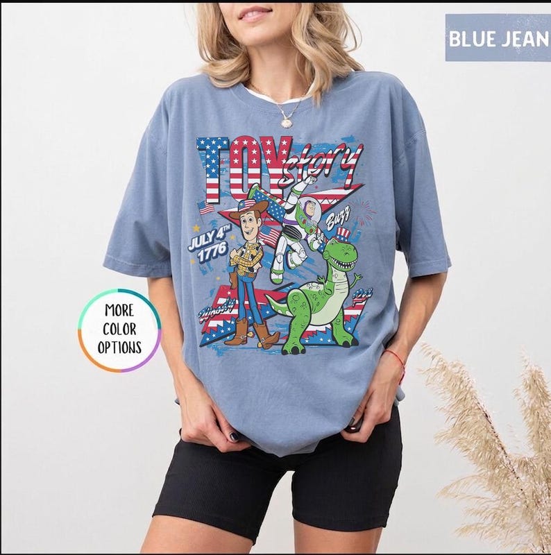 Op de afbeelding: Lichtblauw T-shirt met een Toy Story-afbeelding. Het toont Woody, Buzz Lightyear en een groene dinosaurus, met de tekst "Toy Story" en "July 4th 1776". Het ontwerp bevat elementen van de Amerikaanse vlag. Korte mouwen.