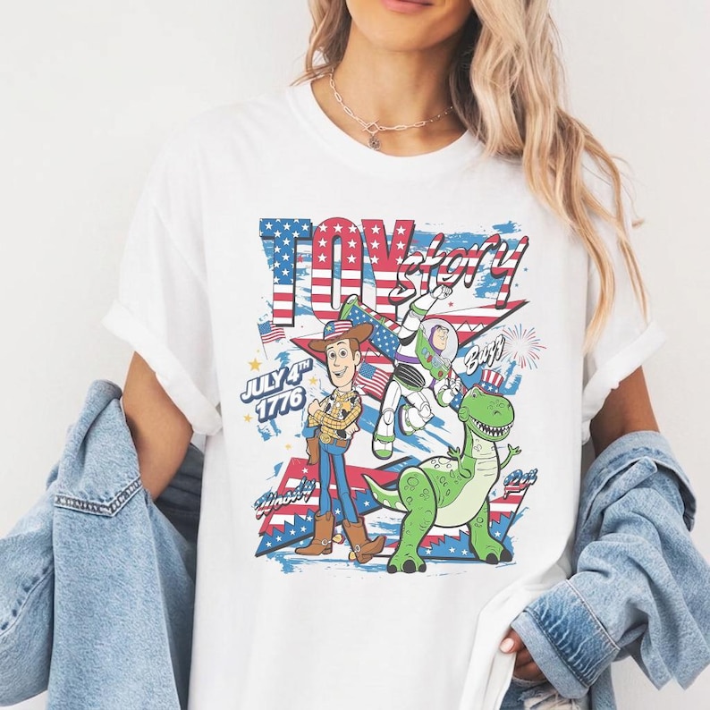 Op de afbeelding: Wit T-shirt met een grafisch ontwerp met de woorden "Toy Story" in rood, wit en blauw. Het ontwerp bevat de stripfiguren Woody, Buzz Lightyear en Rex, met de tekst "July 4th 1776".