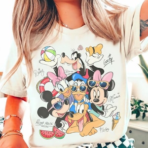 Könnte beinhalten: Cremefarbenes T-Shirt mit einer farbenfrohen Grafik von Disney-Charakteren, darunter Mickey Mouse, Minnie Mouse, Donald Duck und Goofy. Das Design enthält Strandelemente wie einen Strandball, Wassermelonenscheiben und eine Eistüte. Die Namen der Charaktere sind in Schrift geschrieben.