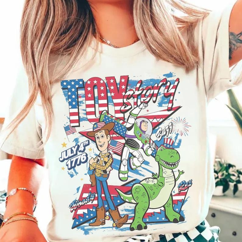 Op de afbeelding: Cr&egrave;mekleurig T-shirt met een patriottisch "Toy Story" ontwerp. De afbeelding bevat Woody, Buzz Lightyear en Rex, met de tekst "Toy Story", "July 4th 1776" en vuurwerk tegen een rood, wit en blauwe Amerikaanse vlag achtergrond.