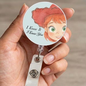 May include: A circular badge reel featuring a light blue background and a watercolor illustration of a person with red hair wearing a red cowboy hat. The text "I Knew It I Knew You" is printed in black script on the left side of the badge reel.