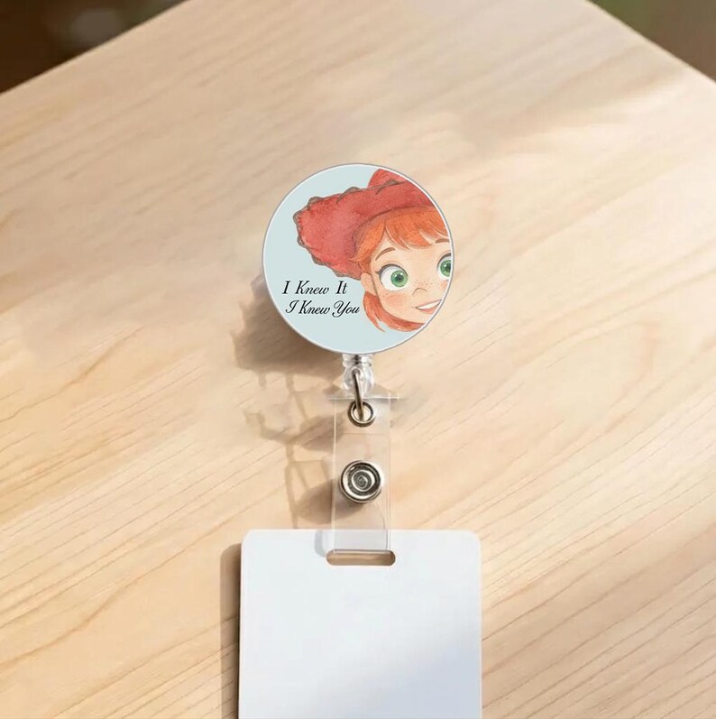 May include: A circular badge reel featuring a light blue background with an illustration of a person with red hair and a red hat. The text "I Knew It I Knew You" is printed in black. The reel has a clear plastic strap and a metal clip.