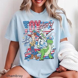 Op de afbeelding: Lichtblauw T-shirt met de tekst "Toy Story" in rood en wit, met een patriottisch ontwerp. Het shirt bevat afbeeldingen van Woody, Buzz Lightyear en Rex, met de tekst "July 4th 1776".