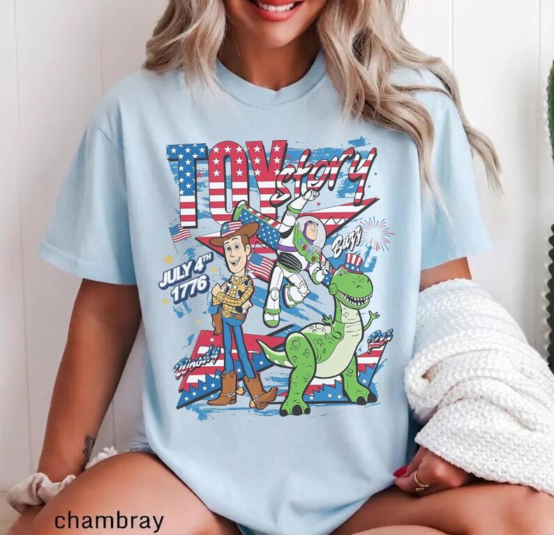 Op de afbeelding: Lichtblauw T-shirt met de tekst "Toy Story" in rood en wit, met een patriottisch ontwerp. Het shirt bevat afbeeldingen van Woody, Buzz Lightyear en Rex, met de tekst "July 4th 1776".