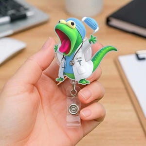 Peut inclure: Un porte-badge de médecin lézard de dessin animé vert et bleu. Le lézard porte une blouse blanche, une blouse bleue et un masque chirurgical. Le porte-badge a un clip en plastique transparent et un cordon rétractable.