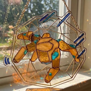 Könnte beinhalten: Buntglas-Suncatcher mit geometrischem Design. Die zentrale Figur ist in Bernstein- und Türkistönen gehalten, mit goldfarbenen Metallkonturen und blauen Akzenten. Es hängt an einer goldenen Kette.