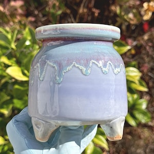 May include: A small ceramic planter with three short legs, featuring a smooth light purple base and a glossy glaze that drips in shades of blue and pink from the rim. The pot has a wide, rounded body and a thick, flat, circular top edge.