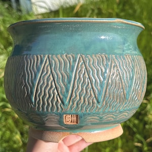 May include: A teal ceramic planter with a textured, wave-like pattern around the middle. The planter has a wide opening and a light brown base. A small, square stamp is visible near the base.