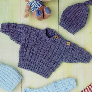 Op de afbeelding: Een set van twee gebreide truien met ruitpatroon, twee bijpassende geribbelde mutsen met kwastjes, een pluche beer met een gestreept shirt en een stoffen zeilbootje met een rood-wit gestreept zeil en een ronde patch met het cijfer twee.