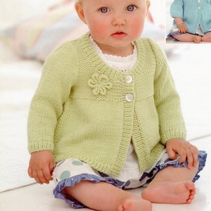May include: A light green knitted cardigan featuring a round neckline, long sleeves, two white buttons, and a small knitted flower detail on the chest. The garment is worn over a white top and white shorts with large blue polka dots and a ruffled hem.