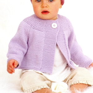 Pode incluir: Um bebê vestindo um casaco de tricô lavanda com um único fecho de botão e um gorro listrado rosa e verde. O bebê está sentado em uma superfície branca e fofa.