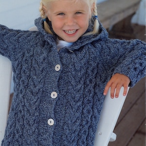 May include: A young girl wearing a blue knitted cardigan with a hood and buttons. The cardigan has a cable knit pattern. The girl is wearing blue jeans.