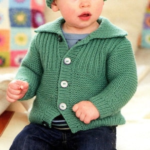 May include: A green knitted cardigan with a button closure and a matching green knitted hat. The cardigan has a collar and is worn by a young child.