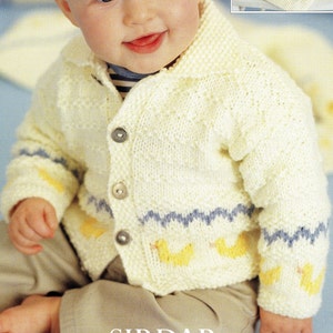 May include: A white knitted cardigan with a yellow and blue pattern. The cardigan has a button fastening and a collar. A baby is wearing the cardigan.