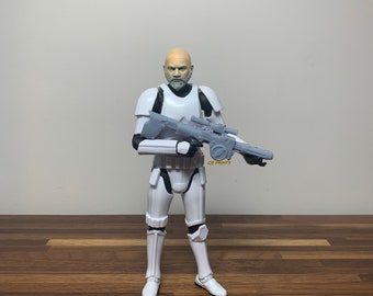 May include: A white action figure of a Star Wars Stormtrooper holding a grey blaster rifle. The Stormtrooper has a realistic face with a beard and grey hair.