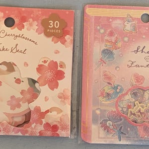 May include: Two packs of decorative stickers. The first pack features a pink and white design with cherry blossoms and birds. The text on the pack reads "Spring Cherryblossoms Flake Seal" and "30 Pieces". The second pack features a pink and blue design with a variety of whimsical illustrations. The text on the pack reads "Shelly Fantasy" and "100 110円(税込)".