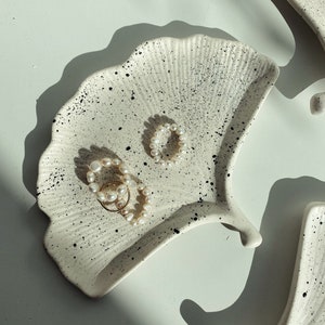 Könnte beinhalten: Zwei weiße Keramikteller in Form von Ginkgoblättern mit schwarzen Flecken. Die Teller liegen auf einer weißen Oberfläche und auf jedem liegt eine Perlenkette.