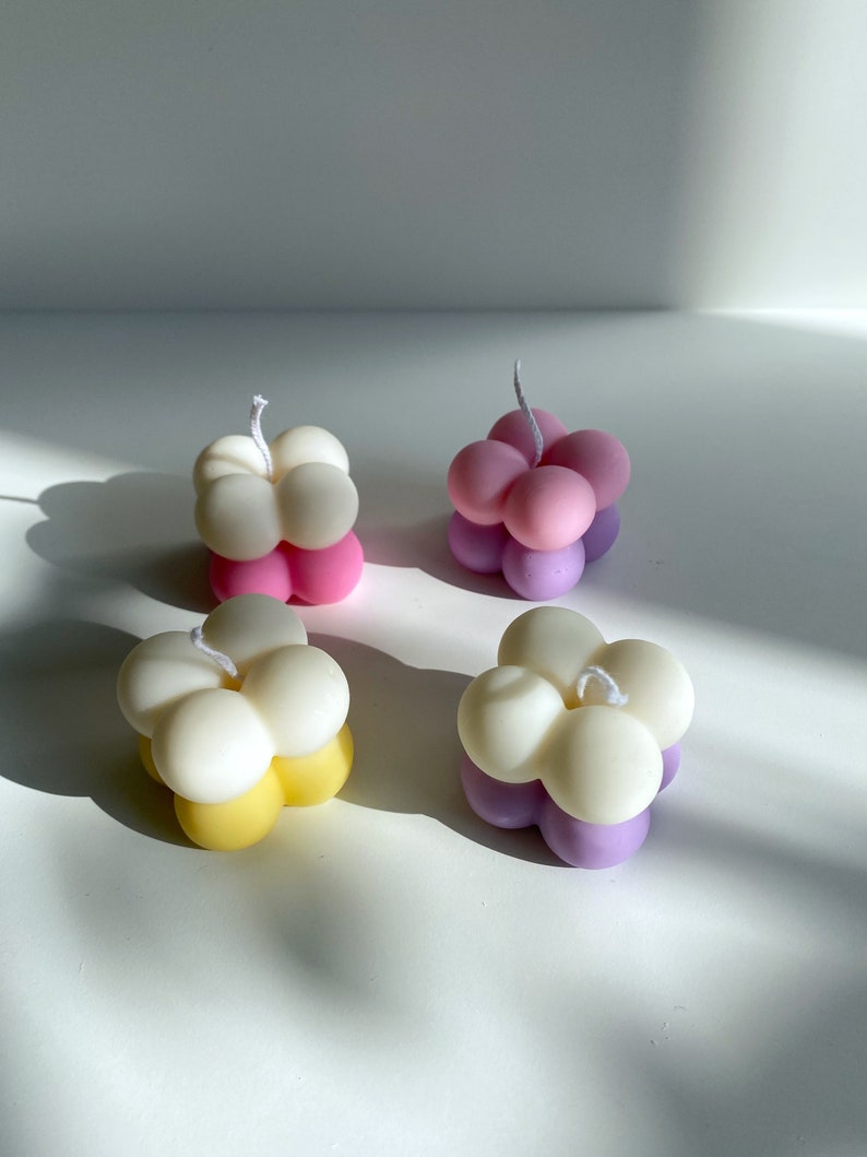 May include: Four small, round, multi-colored candles with a flower-like shape. The candles are white, pink, yellow, and purple.