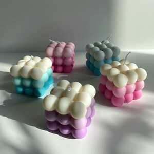 May include: Four square-shaped candles with a bubbled texture. The candles are layered in three colors: white, light blue, and pink. The candles are arranged on a white surface.