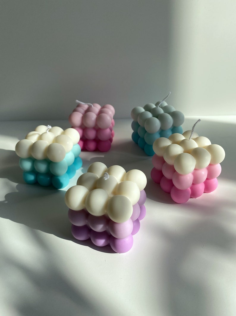 May include: Four square-shaped candles with a bubbled texture. The candles are layered in three colors: white, light blue, and pink. The candles are arranged on a white surface.