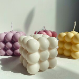 May include: Three square-shaped candles with a bubbled texture. The candles are in shades of purple, white, and yellow.