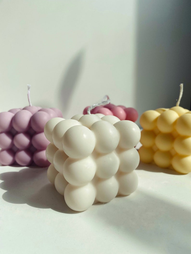 May include: Three square-shaped candles with a bubbled texture. The candles are in shades of purple, white, and yellow.