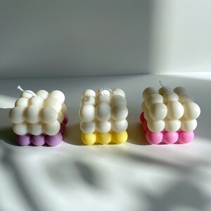 May include: Three square-shaped candles with a bubbled texture. The candles are white with a bottom layer of pink, yellow, and purple.