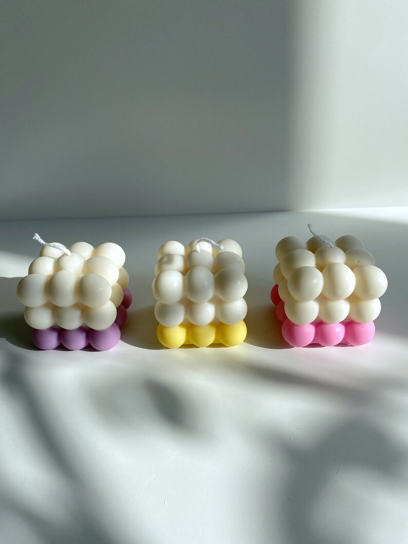 May include: Three square-shaped candles with a bubbled texture. The candles are white with a bottom layer of pink, yellow, and purple.