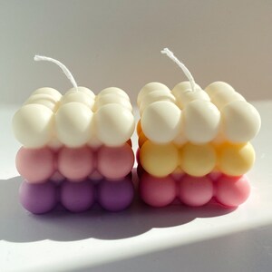 May include: Two square-shaped candles with a bubbled texture. The candles are layered in three colors: white, pink, and purple. The candle on the left is white, pink, and purple. The candle on the right is yellow, pink, and purple.
