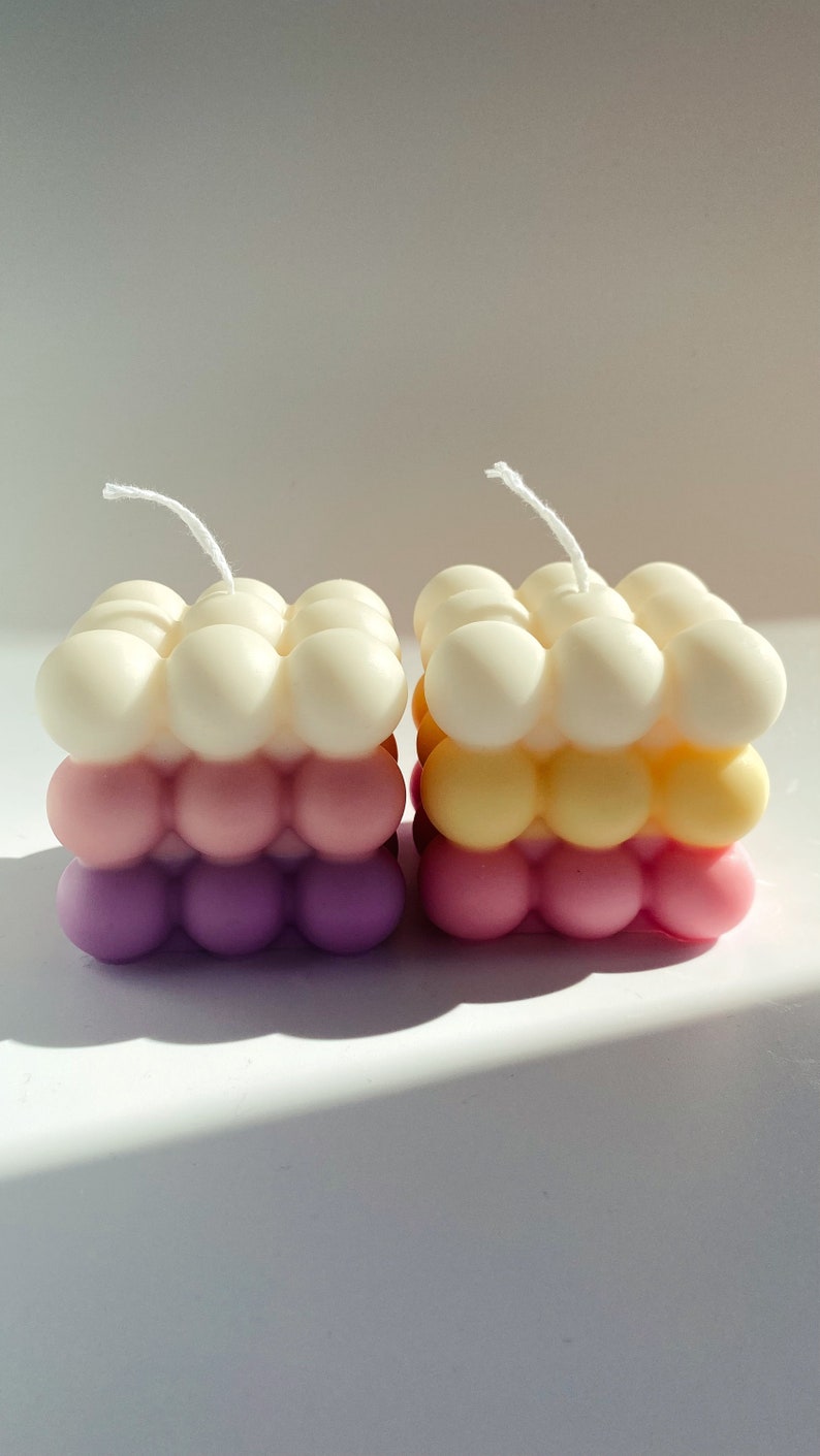 May include: Two square-shaped candles with a bubbled texture. The candles are layered in three colors: white, pink, and purple. The candle on the left is white, pink, and purple. The candle on the right is yellow, pink, and purple.