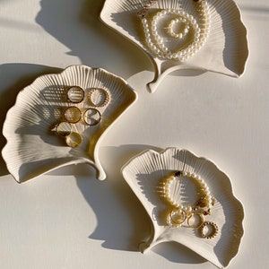 Könnte beinhalten: Drei weiße Keramik-Ginkgoblatt-Schalen mit goldenen Ringen und Perlenketten. Die Schalen stehen auf einer weißen Oberfläche.