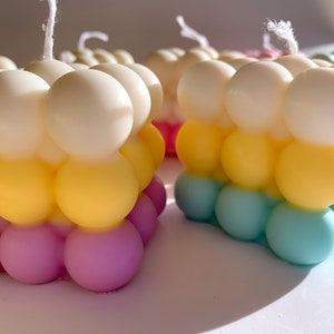 May include: Three colorful bubble candles with white wicks. The candles are layered in white, yellow, and light blue. The bottom layer is a light purple color.