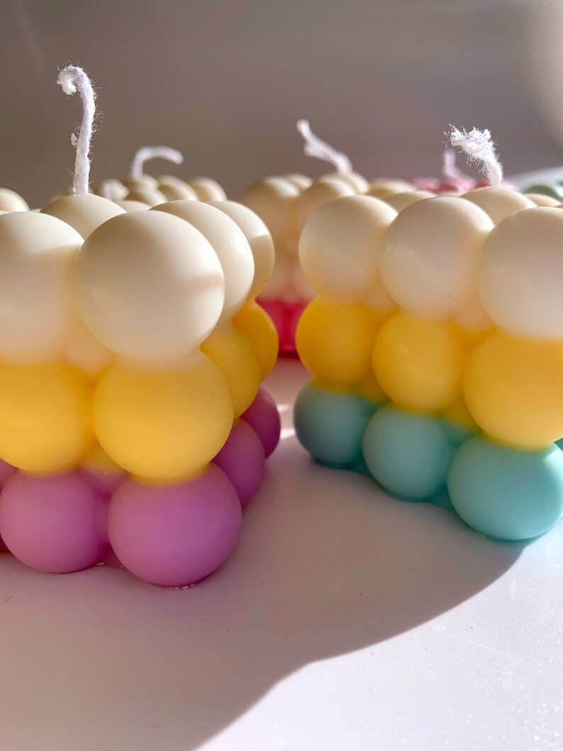 May include: Three colorful bubble candles with white wicks. The candles are layered in white, yellow, and light blue. The bottom layer is a light purple color.