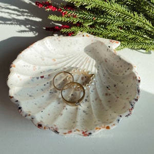 Może przedstawiać: Biała ceramiczna miseczka w kształcie muszli z wzorami w odcieniach pomarańczowego, brązowego i czarnego. Miseczka jest wypełniona złotymi pierścionkami i naszyjnikiem z pereł.