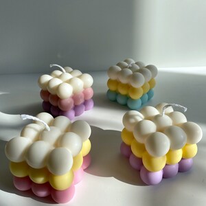 May include: Four square-shaped candles with a bubbled texture. The candles are layered in three colors: white, yellow, and pink.