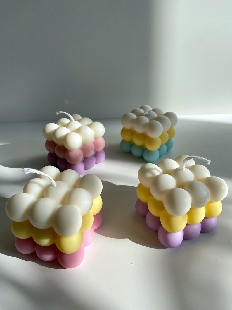 May include: Four square-shaped candles with a bubbled texture. The candles are layered in three colors: white, yellow, and pink.