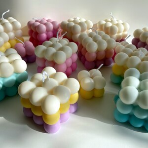 May include: A collection of colorful, square-shaped candles with a unique bubbled texture. The candles are layered in shades of pink, yellow, blue, and white.