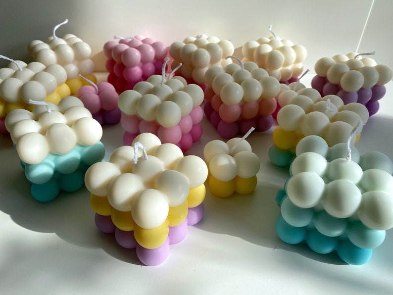 May include: A collection of colorful, square-shaped candles with a unique bubbled texture. The candles are layered in shades of pink, yellow, blue, and white.