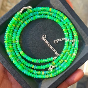 May include: A necklace composed of small, round, vibrant green opal beads with a reflective, iridescent quality. The strand features a silver-toned metal lobster claw clasp and an adjustable chain extender, creating a circular arrangement.